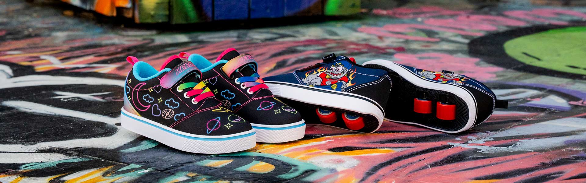 Two pairs of black kids Heelys roller shoes with colorful designs on a graffiti-covered surface